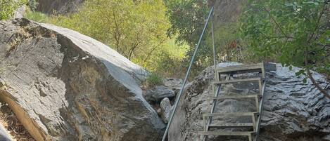 Ein felsiges Gelände mit einer Metalltreppe, die einen steilen Hang hinaufführt, umgeben von großen Felsen und spärlicher Vegetation.