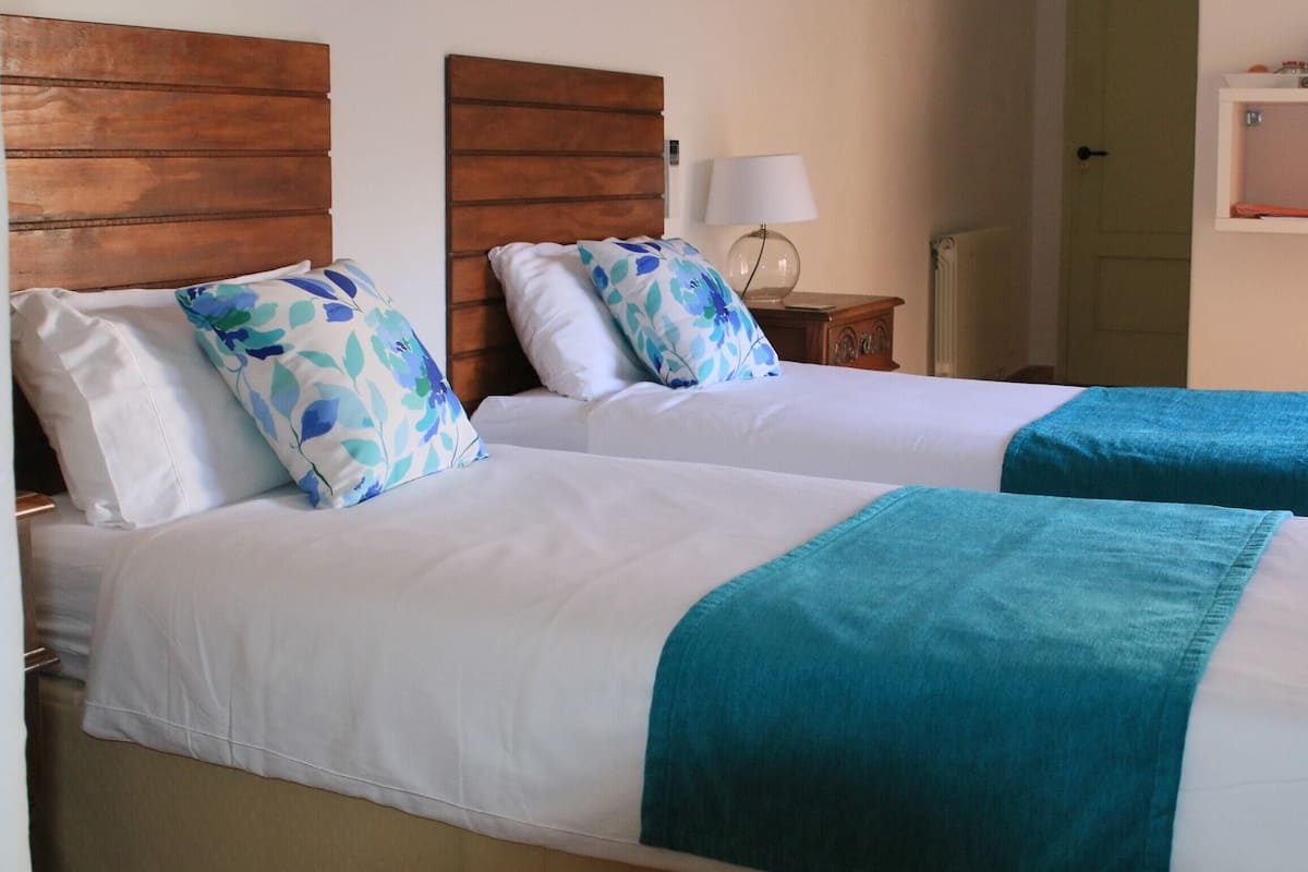 Two beds with white linens and teal blankets, wooden headboards, a bedside table with a lamp, and a shelf with various items.