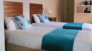 Two beds with white linens and teal blankets, wooden headboards, a bedside table with a lamp, and a shelf with various items. - Casa del Patio PdV (El Pinar)