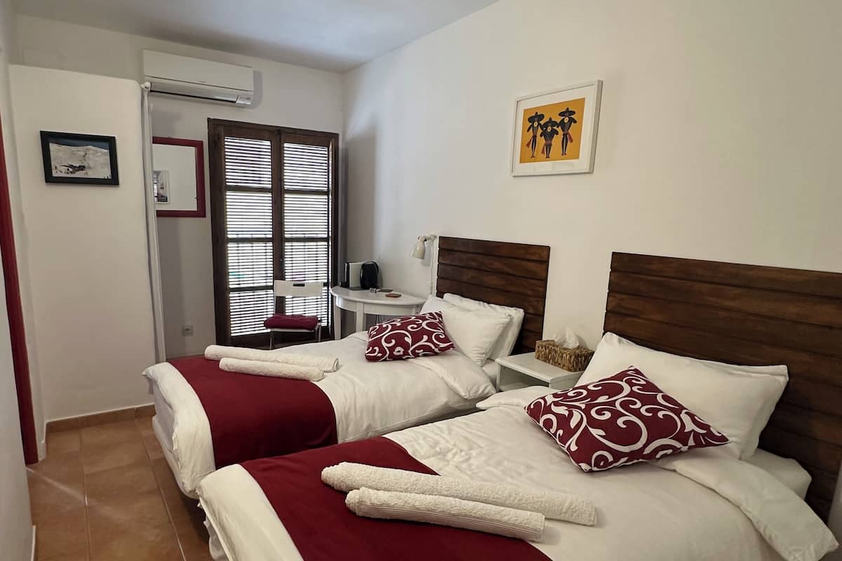 A hotel room with two beds, each with white and red bedding, a small table with a basket, and a window with blinds.