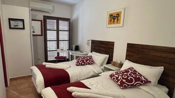 A hotel room with two beds, each with white and red bedding, a small table with a basket, and a window with blinds.