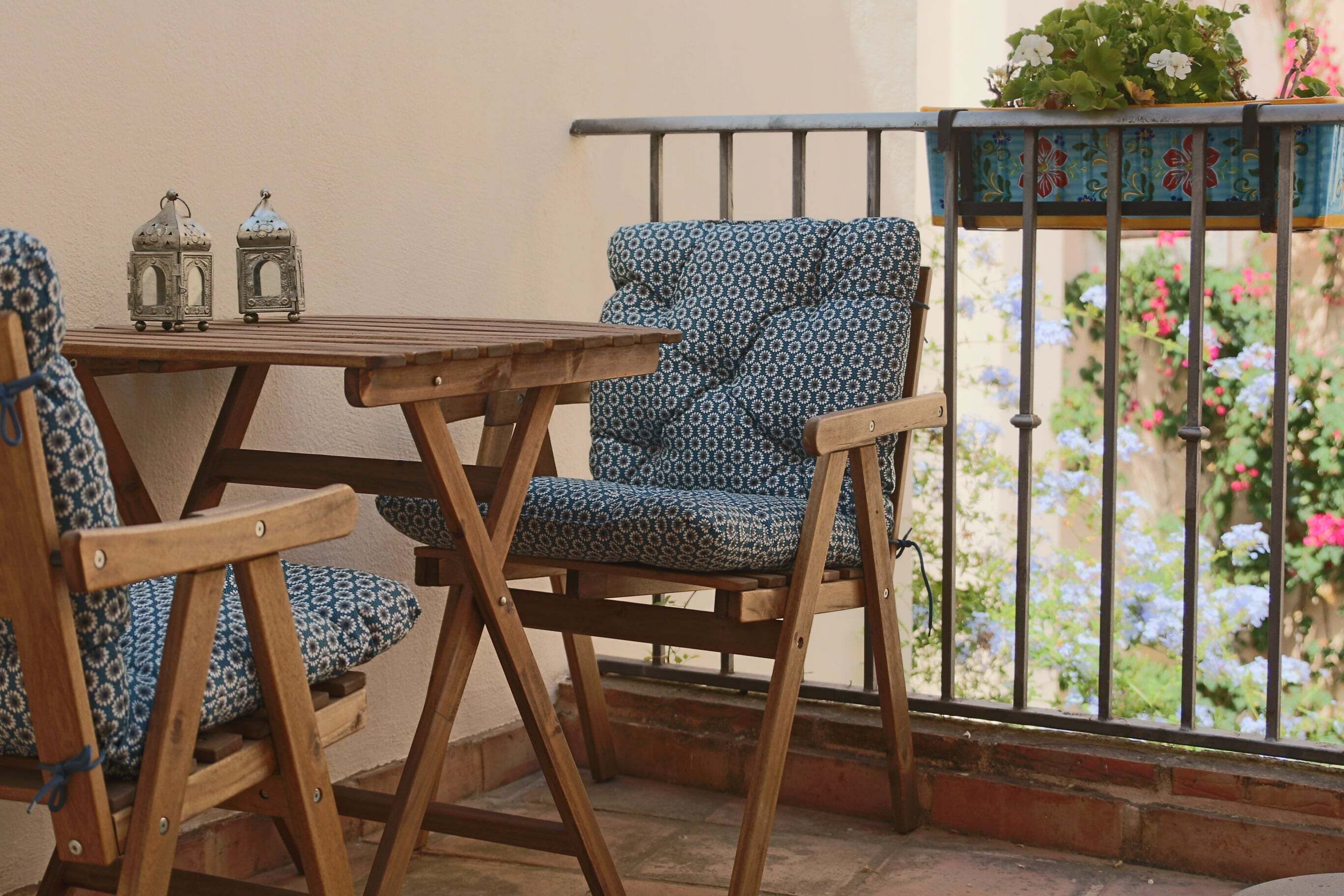 Una mesa de madera y dos sillas con estampado, junto a una baranda de metal y plantas en macetas al fondo.