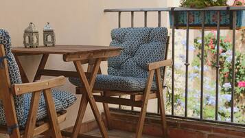 Una mesa de madera y dos sillas con estampado, junto a una baranda de metal y plantas en macetas al fondo.