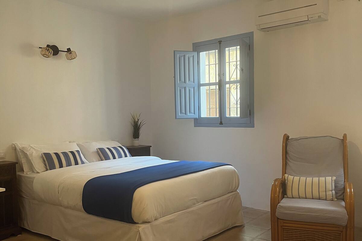 A bedroom with a bed, a chair, a window, and a hanging light fixture.