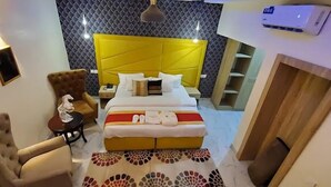 Hotelkamer met een geel hoofdeinde, een bed met witte lakens, een klein rond tafeltje met een lamp, een stoel en een kamerkleed met dessin.