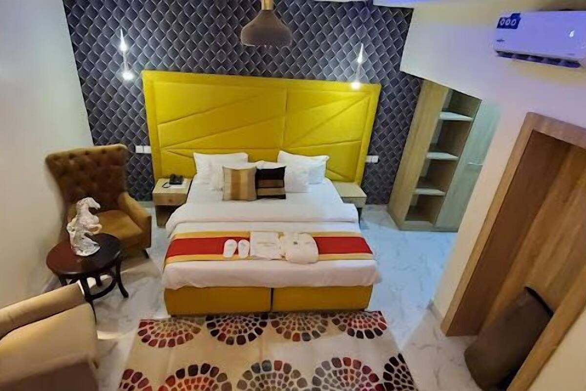 Quarto de hotel com cabeceira amarela, cama com roupa de cama branca, mesa redonda pequena com abajur, uma cadeira e tapete estampado.