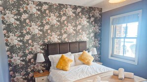 A bedroom with a floral wallpaper, a bed with white linens and two pillows, a nightstand with a lamp, and a window with a view. - Dingle Town Holiday Home (Dingle)