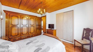 Un dormitorio con un armario grande de madera, una cama con colcha estampada con hojas, un televisor sobre un mueble de madera y una silla de madera.