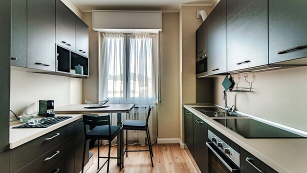 A modern kitchen with dark cabinets, a built-in oven, and a dining area with a table and chairs. - Turchese (Como)