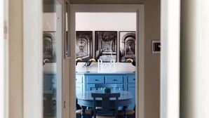 A hallway leading to a kitchen with a blue cabinet, a round table, and chairs. There are three framed pictures on the wall above the cabinet. - Turchese (Como)