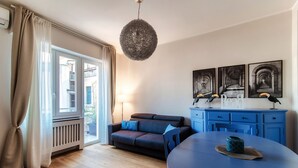 A modern living room with a sofa, a dining table, and a kitchen area with blue cabinets and a hanging light fixture. - Turchese (Como)