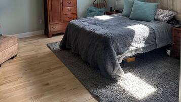 Un dormitori amb llit, una petita arca de fusta, ventilador de sostre i dues finestres amb cortines estampades.