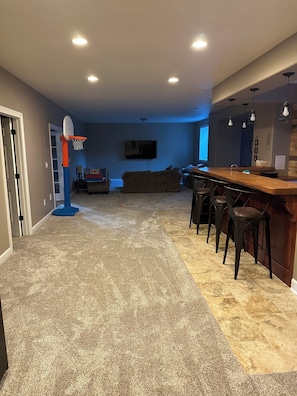 A home interior with a basketball hoop, a bar area with stools, a large TV, and a carpeted floor. -  4 bedroom house 7 min.to Erin Hills SAND VALLEY GOLF COURSE HOLY HILL WHISTLING (Hartford)