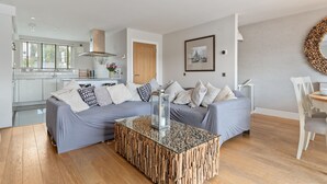 A modern living room with a glass coffee table, grey sofa, and a dining area with a white chair. - Ger Y Mor - Village Centre With Parking (Saundersfoot)