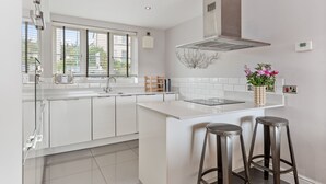 A modern kitchen with white cabinetry, a central island with stools, a stainless steel range hood, and a window with blinds. - Ger Y Mor - Village Centre With Parking (Saundersfoot)
