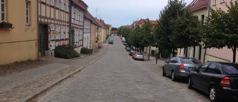 A cobblestone street lined with traditional half-timbered houses on one side and a row of parked cars on the other.