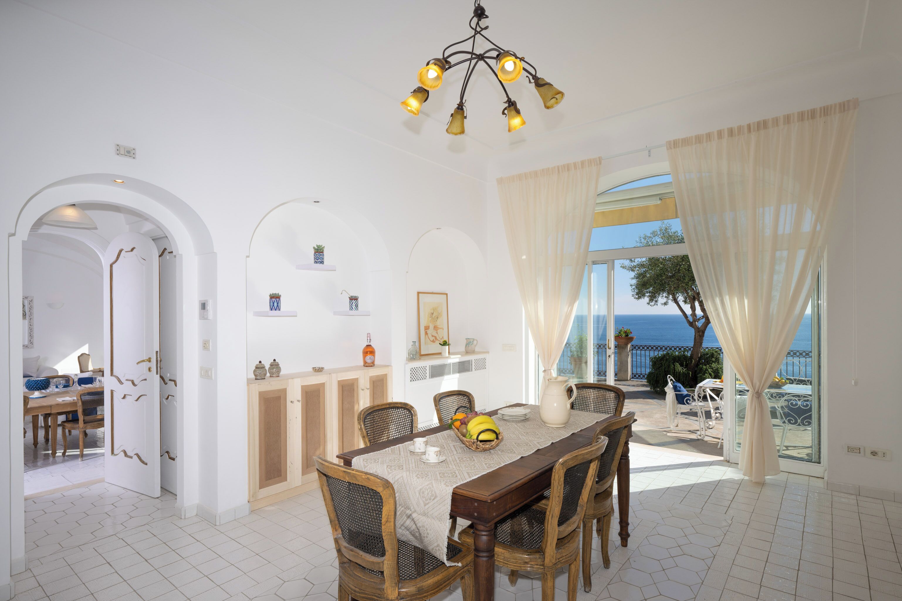 A dining area with a table set for six, a chandelier, and a view of the sea through large windows.