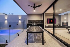 Een moderne eethoek buiten met een zwembad, een plafondventilator, een keuken met grill en een televisiescherm.