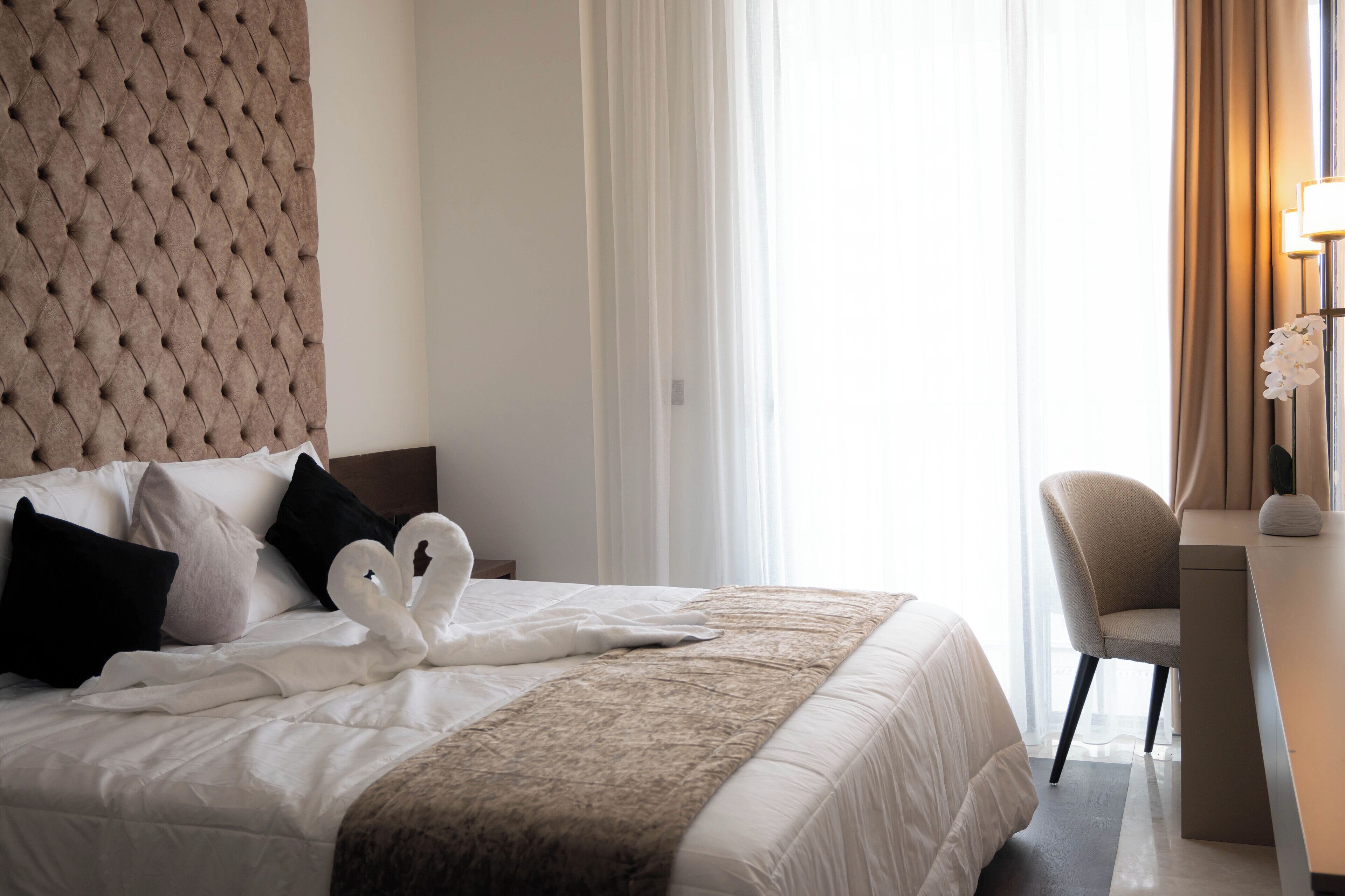 Una habitación de hotel con cabecera capitonada, cama con ropa de cama blanca, dos almohadas con forma de cisne, un escritorio con silla y una lámpara.