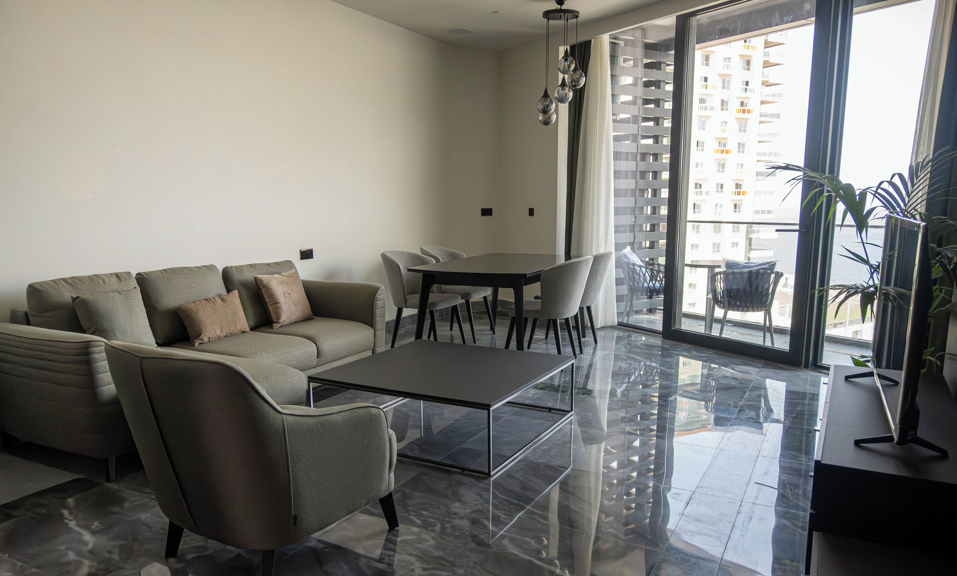 A modern living room with a sofa, a coffee table, and a dining table with chairs. There is a large window with a view of a building.