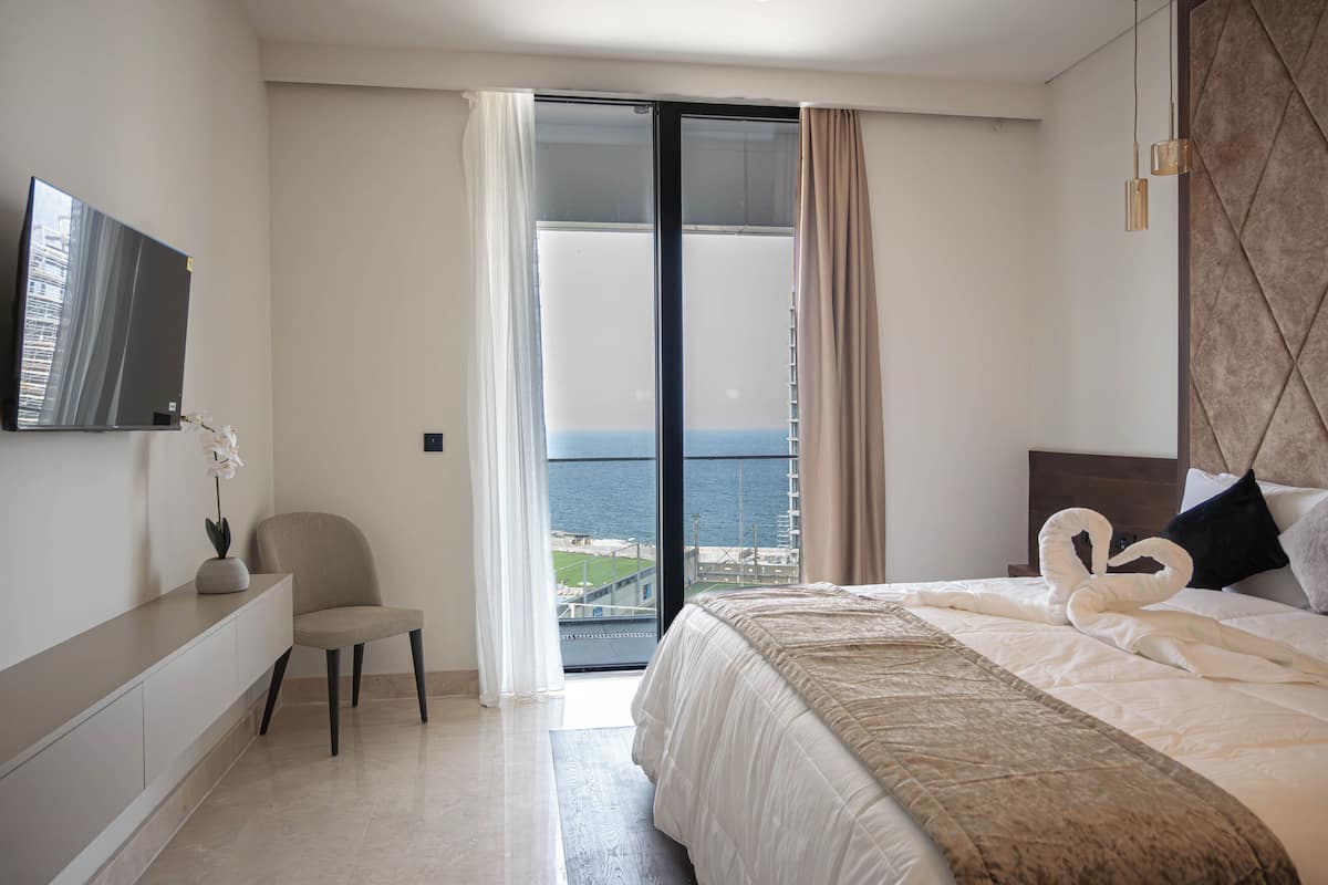 Habitación de hotel moderna con cama, una silla, televisor y vista al mar a través de un ventanal grande.
