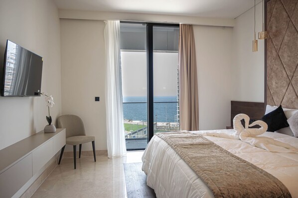 Habitación de hotel moderna con cama, una silla, televisor y vista al mar a través de un ventanal grande.