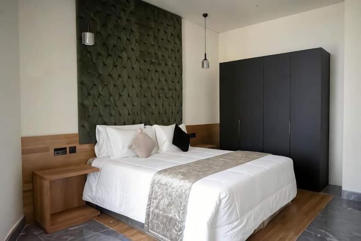 Habitación de hotel moderna con una cama grande, una mesita de noche de madera, un armario oscuro y una pared verde con textura.