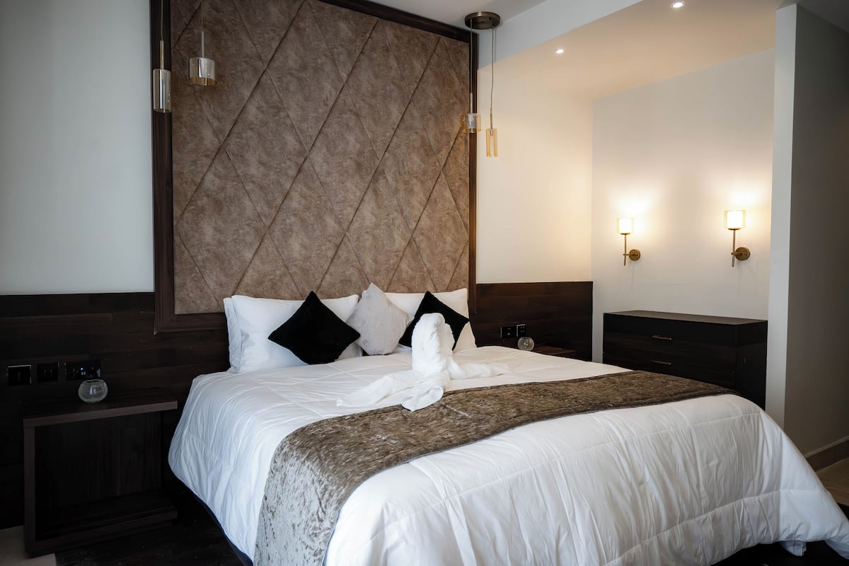 Habitación de hotel con una cama grande, cabecera de madera, mesita de noche y dos lámparas de pared.