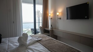 A hotel room with a bed, a desk, a chair, a TV, and a view of the sea. - Best View Fortina Luxury Suites  (Sliema)