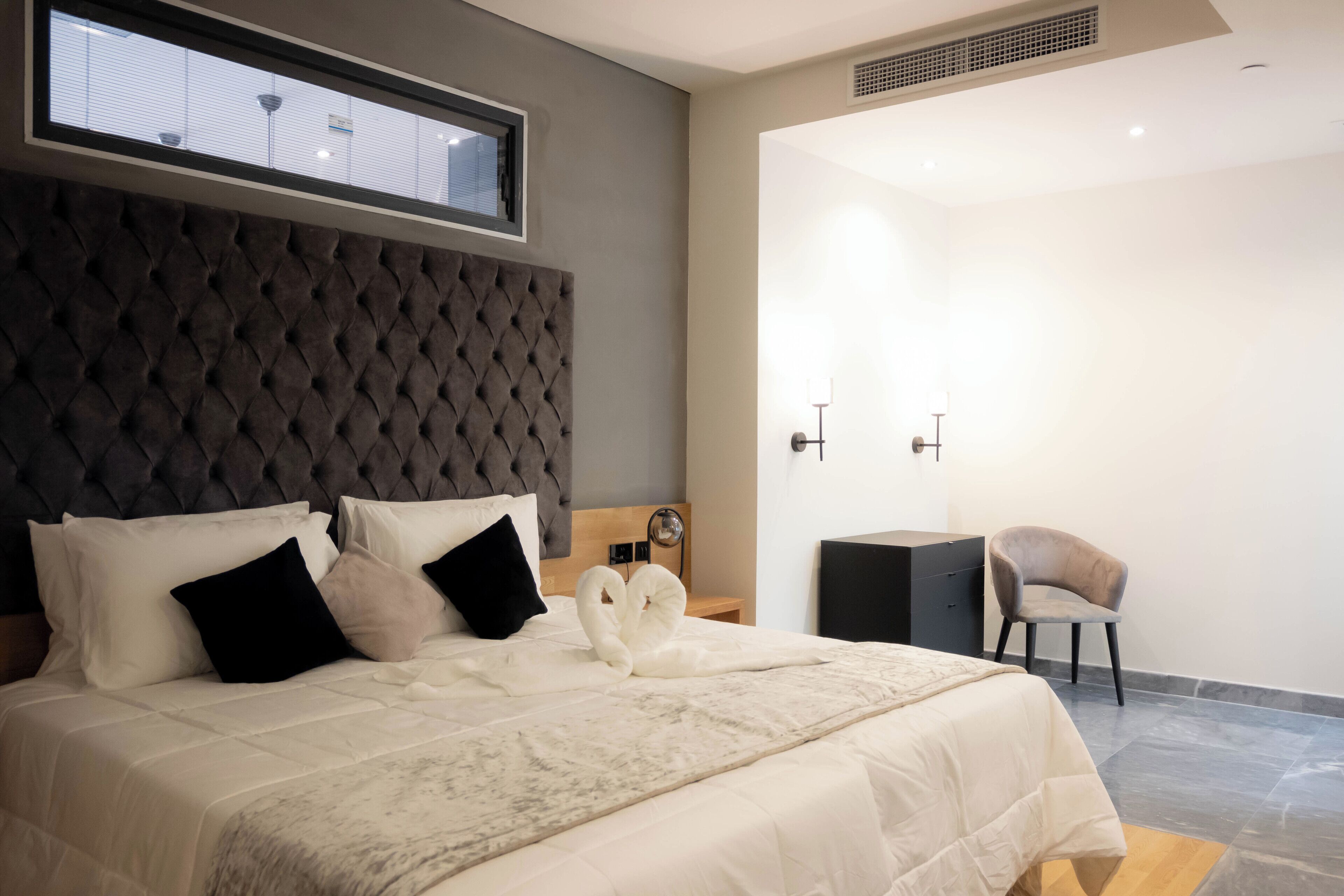 Un dormitorio moderno con un cabecero capitonado, una cama bien tendida con ropa blanca, una mesita de noche con una lámpara y una silla.