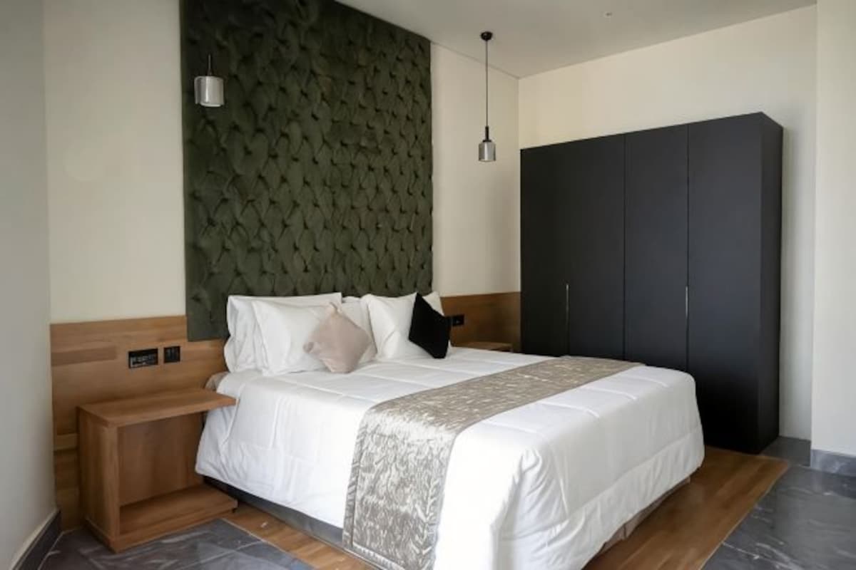 A modern hotel room with a large bed, a wooden nightstand, a dark wardrobe, and a green textured wall.