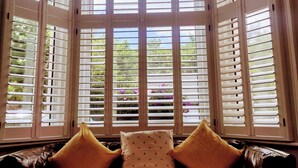 A room with a bay window featuring white shutters, a leather sofa with two yellow cushions, and a view of greenery and flowers outside. - Annette's Place, Greenwich (London) - Garden Apartment (close to the O2 Arena) (London)