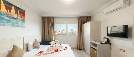 Ein Hotelzimmer mit Bett, einem Gemälde, einem Fernseher, einer Minibar und Blick auf Pyramiden.