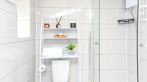 A modern bathroom with white tiles, a glass shower enclosure, a white toilet, and a shelf with toiletries and a small plant.