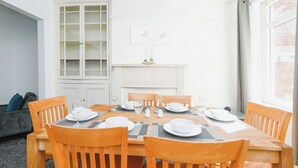 A dining area with a wooden table set for four, surrounded by wooden chairs. A white cabinet with glass doors is to the left, and a fireplace with a small plant on the mantel is in the background.