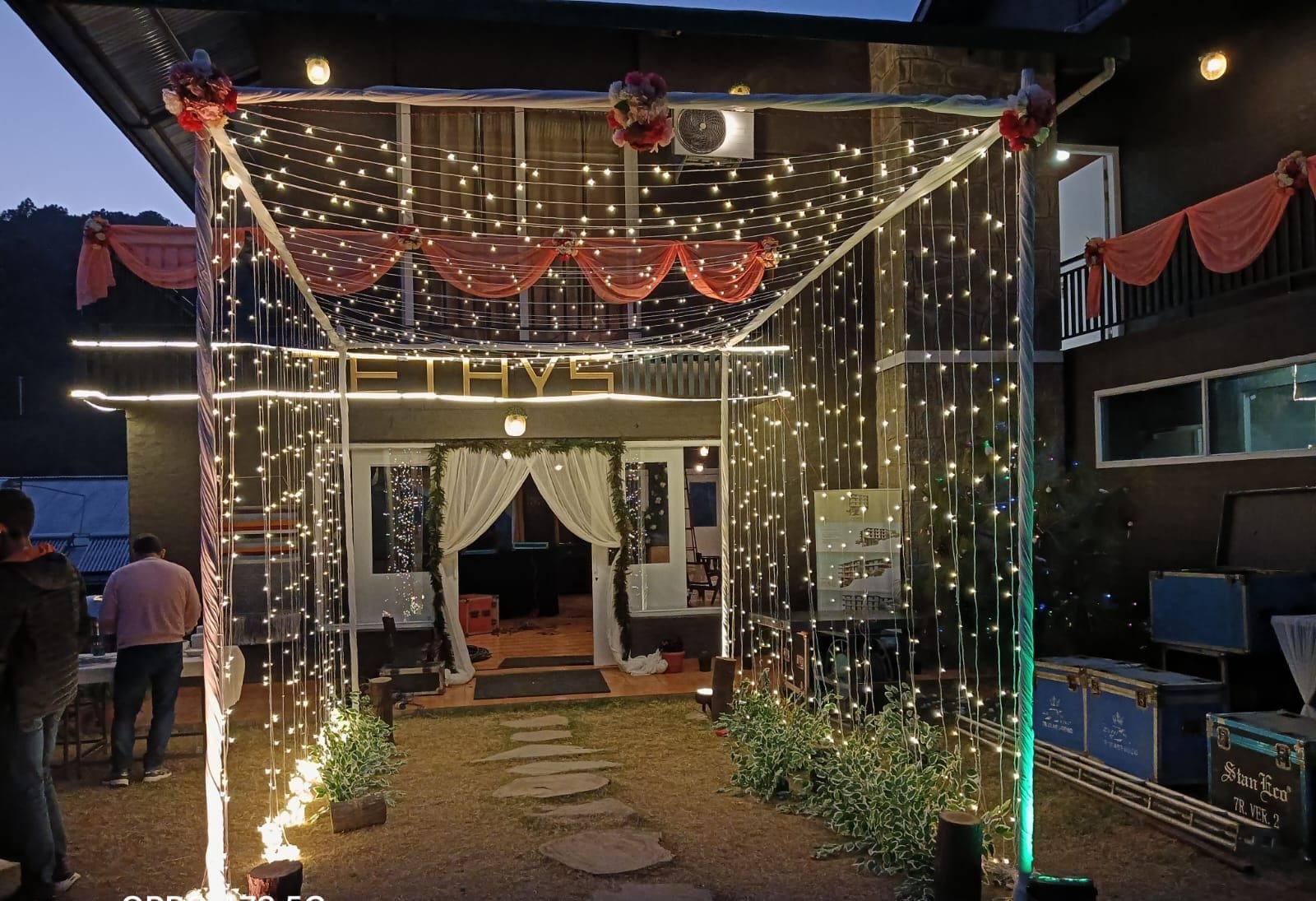 Un espace extérieur aménagé avec des guirlandes lumineuses, des étoffes suspendues et un chemin menant à l’entrée d’un bâtiment.