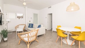 Una sala moderna con un sofá blanco, sillones de mimbre, una mesa de comedor con sillas amarillas, un ventilador de techo y una lámpara colgante.