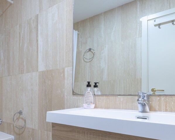 Une salle de bain carrelée de marbre beige, avec un lavabo blanc et un miroir, un porte-serviettes et un flacon de savon pour les mains.