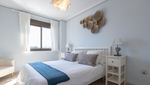 A bedroom with a large bed, a bedside table with a lamp, a chair, and a wall art piece. - Pacifico Playa in M Laga (Málaga)