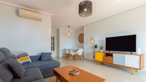 A modern living room with a grey sofa, a wooden coffee table, a TV on a wooden stand, and a dining area with a table and chairs. - Pacifico Playa in M Laga (Málaga)