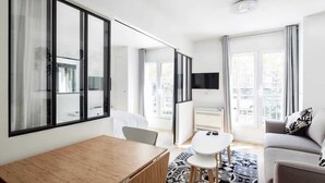 A modern hotel room with a wooden desk, a small round table, a grey sofa, and a TV on the wall. - Résidence Paris Rochechouart (Paris)