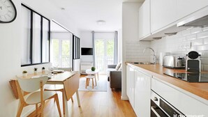 A modern kitchen with white cabinets, a wooden countertop, and a built-in oven. There is a dining area with a table and chairs, and a large window providing natural light. - Résidence Paris Rochechouart (Paris)
