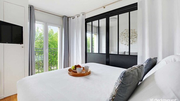 A modern bedroom with a large bed, a flat-screen TV mounted on the wall, and a balcony with a view of greenery. - Résidence Paris Rochechouart (Paris)