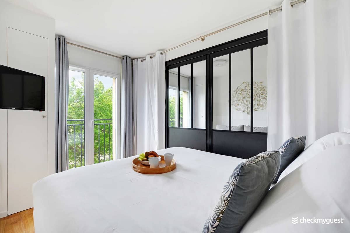 Une chambre moderne avec un grand lit, un téléviseur à écran plat fixé au mur et un balcon offrant une vue sur la verdure.