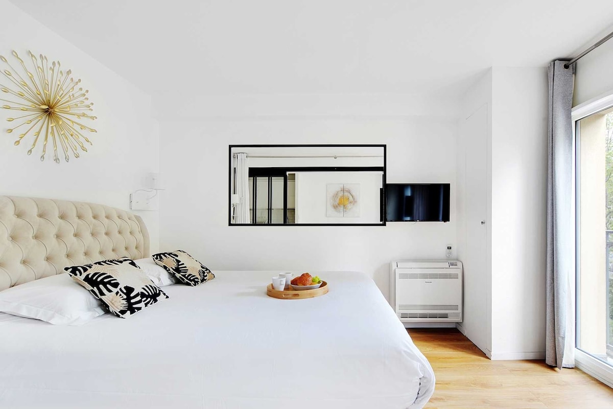 A modern bedroom with a tufted headboard, a bed with patterned pillows, a bedside table with a bowl of fruit, a flat-screen TV, and a large window with a view of greenery.