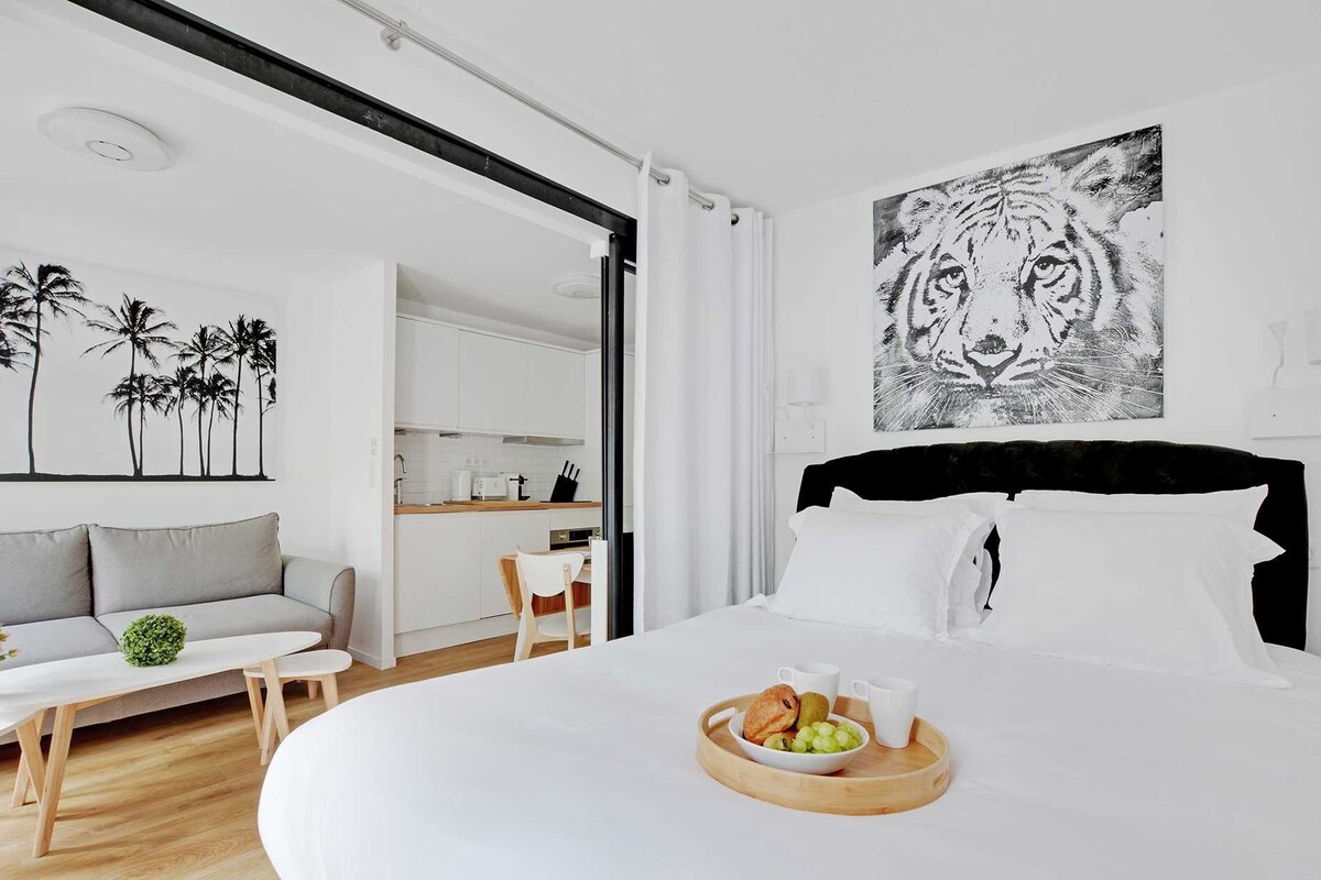 A modern hotel room with a large bed, a dining area, and a kitchen visible through an open door.