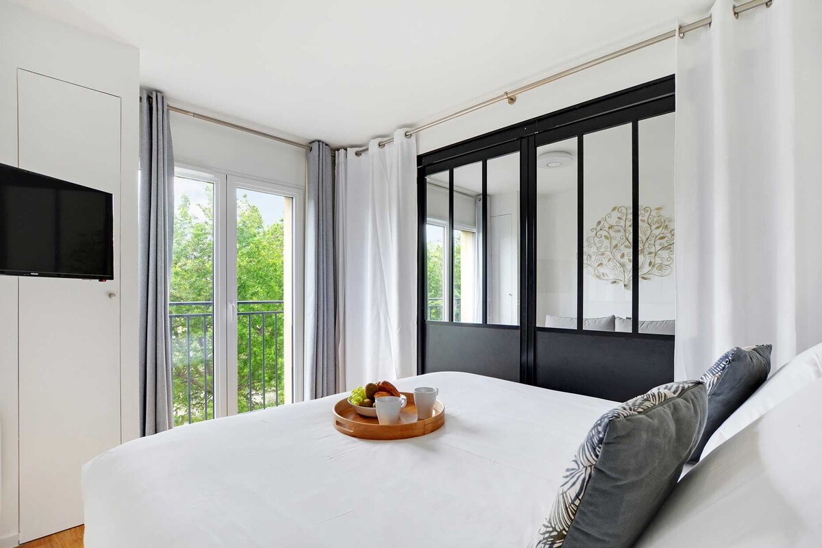 A modern bedroom with a large bed, a flat-screen TV mounted on the wall, and a balcony with a view of greenery.