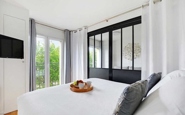 A modern bedroom with a large bed, a flat-screen TV mounted on the wall, and a balcony with a view of greenery. - Résidence Paris Rochechouart (Paris)