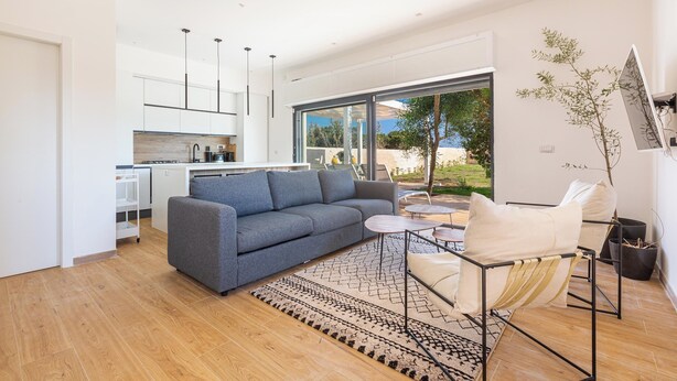 A modern living room with a grey sofa, a coffee table, and a chair. There is a kitchen in the background with white cabinets and a large window. - 4147 Villa Daversa by Barbarhouse (Manduria)