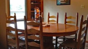 Een eetgedeelte met een ronde houten tafel, omringd door houten stoelen. Er is een raam met gordijnen, een houten kast met decoratieve objecten, en een lijst met een foto aan de muur.